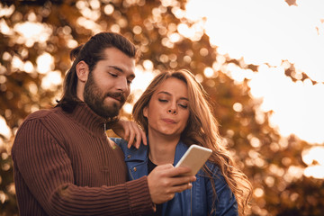 Young couple using cellphone in autumn colored park.