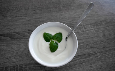 Ceramic bowl with white plain yogurt, mint leaves and spoon isolated on dark wooden background from above