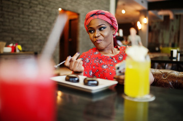 Stylish african woman in red shirt and hat posed indoor cafe, eat chocolate dessert cakes and looking at mobile phone.