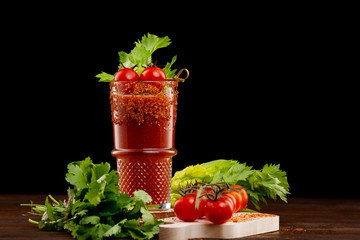 Glass of bloody mary cocktail decorated with celery and pepper and a branch of red tomatoes on a wooden table isolated at black background.