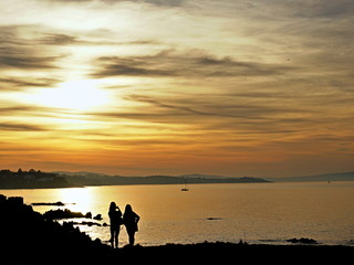 Fototapeta na wymiar Summer memories. Silhouettes of two girls taking photo of sunset over The Irish Sea