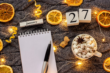 Winter composition. Wooden calendar January 27th Cup of cocoa with marshmallow, empty open notepad with pen, dried oranges, light garland on grey knitted background. Top view Flat lay Mockup