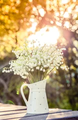 Draagtas Bouquet of white flowers Lily of the valley (Convallaria majalis) in a white dotted jug shaped vase, outdoors on a table, trees on background beautiful golden hour light. © FotoHelin
