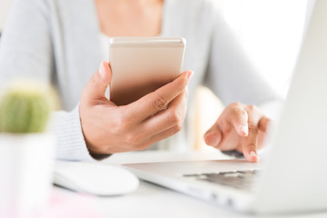 Woman hand holding white mobile phone on a table with a laptop in office.