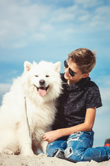Happy 11 year old boy playing with his dog breed Samoyed at the seashore against a blue sky close up. Best friends rest and have fun on vacation, play in the sand