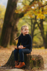 A little girl in black clothes is sitting on a stump and playing with autumn leaves. Blond hair girl