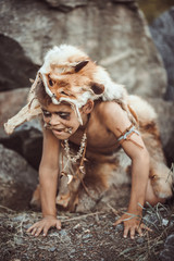 Fototapeta na wymiar Caveman, manly boy hunting outdoors. Prehistoric tribal boy outdoors on nature. Young shaggy and dirty savage, warrior and hunter hiding in an ambush behind a stone in cave. Primitive ice age man in