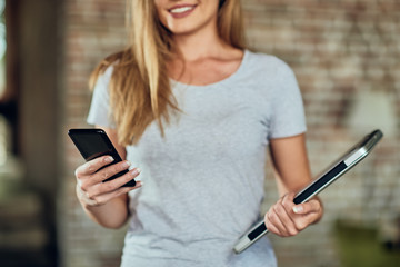 Close up of woman holding laptop and using smart phone for texting. In background brick wall.