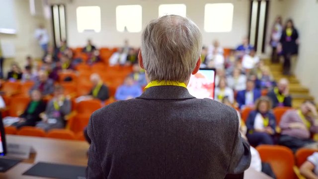 elderly professor is speaking in front of audience of scientists in conference, people are listening