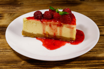 Piece of tasty New York cheesecake with raspberries and raspberry jam in a white plate on wooden table