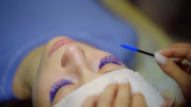 lashmaker is brushing extended blue lashes by cosmetic brush, client is lying on couch, close-up