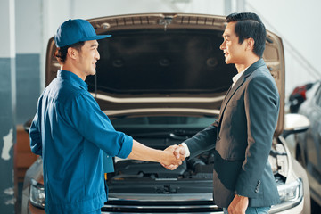 Asian young auto mechanic in uniform and a client are shaking hands and smiling with car in the...