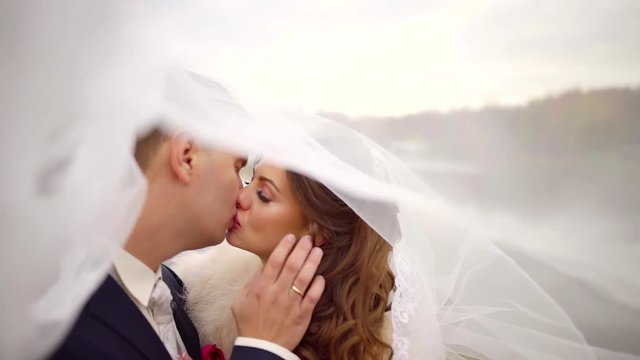 happy groom and bride are kissing tenderly under white wedding veil, man is touching woman