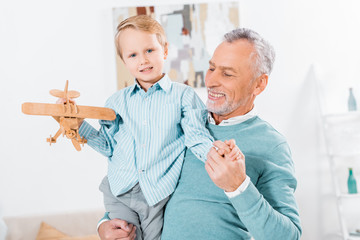 selective focus of smiling middle aged man holding little grandson with wooden airplane on hands at home