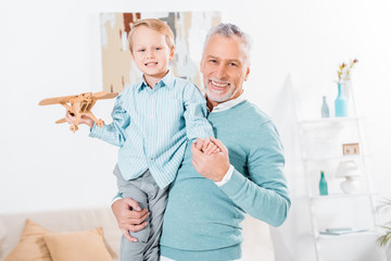 happy middle aged man holding little grandson with wooden airplane on hands at home