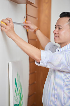 Young man measuring the wall with tape measure and trying to hang the picture