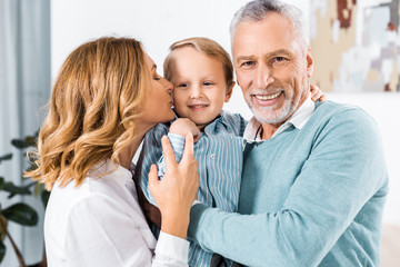 selective focus of cheerful middle aged couple holding adorable grandson and kissing him in cheeks