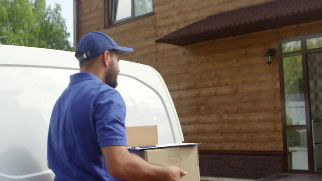 Handheld tracking shot of male courier in blue uniform taking packages out of van and delivering it to happy blond woman standing on doorstep. She is smiling and signing receipt on clipboard