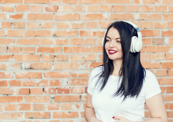 Portrait of a smiling brunette girl on the brick wall background. Listening to music in white headphones. Youth culture, album cover.