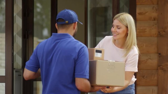 Medium shot of male courier in blue uniform delivering boxes to happy blond woman. She is signing receipt on clipboard and smiling