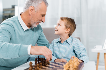 side view of happy grandson and grandfather looking at each other while playing chess at home