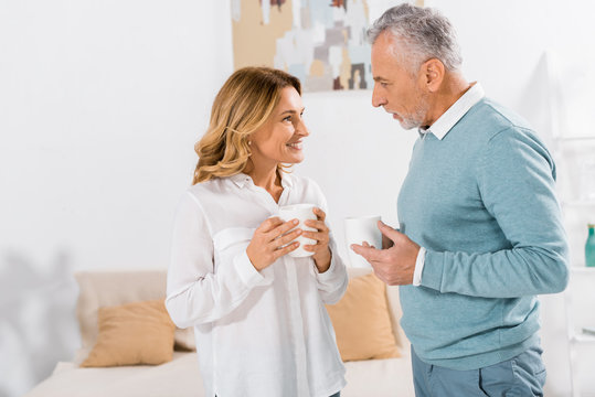 side view of happy woman with coffee cup talking to mature husband at home