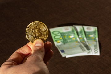 Hand hält Bitcoin Münze mit Hundert Euro Scheinen im Hintergrund