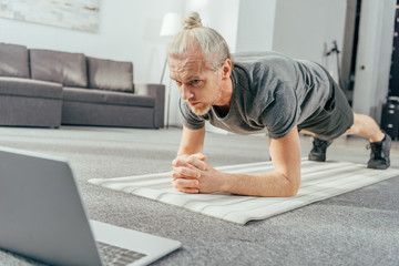 sporty adult man doing plank exercise and looking at laptop