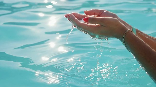 Splendid view of two woman`s hands raising sparkling water drops over a wading pool outdoors with beautiful turquiose waters on sunset in summer