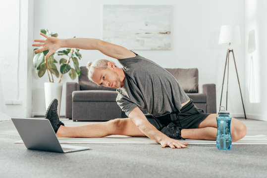 sporty adult man stretching on yoga mat and looking at laptop
