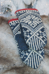 Traditional knitted warm mittens with a white and blue pattern are on a fur cover against a snowy Christmas tree with festive lights. Concept: Merry Christmas and Happy New Year holidays. Vertical. 