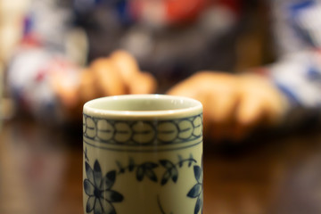 White ceramic cup of Japanese roasted green tea on a table in traditional Japanese restaurant, with young asian woman using smartphone and waiting for her food in the background (The tea is called Hoj