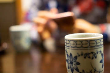 White ceramic cup of Japanese roasted green tea on a table in traditional Japanese restaurant, with young asian woman using smartphone and waiting for her food in the background (The tea is called Hoj
