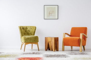 Olive green stylish armchair next to wooden coffee table and orange vintage armchair in tasteful living room interior with camel poster on white wall