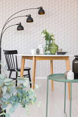 Elegant dining room with round table with vases and flowers next to black wooden chair and industrial metal lamp, real photo with unique wallpaper