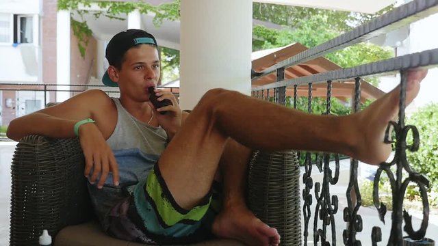  Cheery view of a young brunette teenager dressed in a striped singlet and shorts sprawling in an armchair with an electronic cigarette in mouth. 