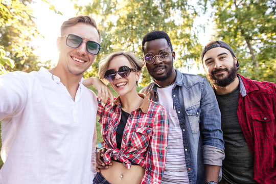 Stylish young people adult men and women using smartphone visit public park during their day off, making a group photo in park. Guys wearing casual clothing and sunglasses. - Powered by Adobe