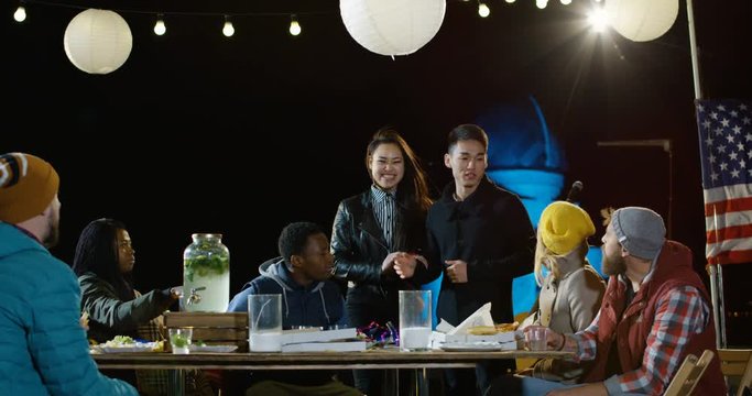 Medium shot of an asian couple at a rooftop party announcing their marriage to their happy friends