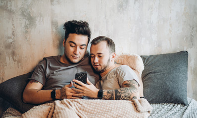 Two contented homosexual male people lying together in bed and using smartphone to make video call to friends