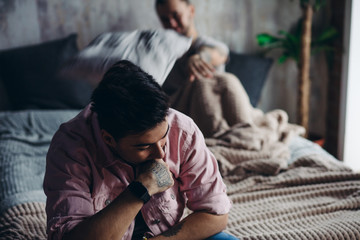Fototapeta premium Young gay couple going through relationship problems. Pensive gloomy man dressed in t-shirt and underwear sits on bed with his hand leaning on chin with blurred image of his bridegroom on background