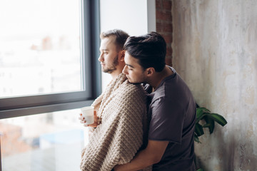 Romantic indoor shot of caucasian gay couple standing against window in modern loft design studio,...