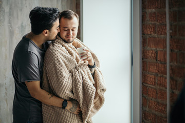 Romantic indoor shot of caucasian gay couple standing against window in modern loft design studio, looking at the street, dominant man taking care of his mate, tucking him with plaid