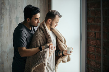 Romantic indoor shot of caucasian gay couple standing against window in modern loft design studio, looking at the street, dominant man taking care of his mate, tucking him with plaid