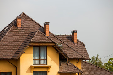 Top of big modern expensive residential house with shingled brown roof, high brick chimneys, stucco walls and plastic attic windows on blue sky copy space background. Real estate selling concept.