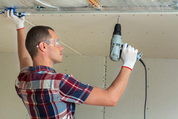 Young man in goggles fixing drywall suspended ceiling to metal frame using electrical screwdriver...
