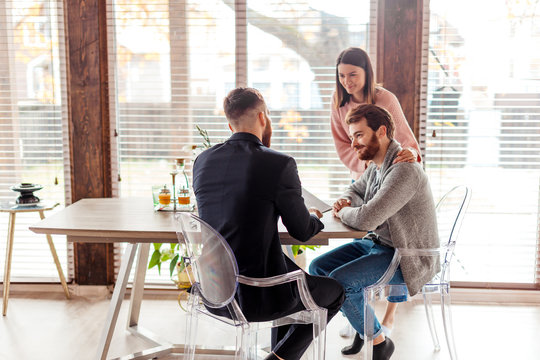 Successful Lawyer Giving Consultation To A Young Attractive Family Couple About In His Modern Office, Buying Renting Apartment Or House.