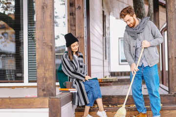 Young rich couple spend a quiet peaceful day outdoor in their country house, relaxing from the urban fuss. Young man sweeps the leaves, clean the garden. Private house purchase concept.