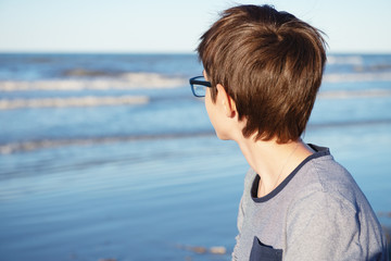 Young boy posing at the summer beach. Cute spectacled 12 years old boy at seaside, looking at sea. Kid's outdoor portrait over seaside.