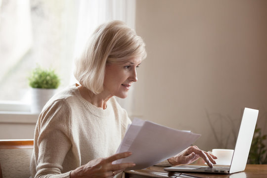 Senior Woman Holding Papers Busy At Laptop Managing House Utility Bills Or Finances, Aged Female Using Computer Working With Bank Loan Or Mortgage Documents Online. Elderly And Technology Concept