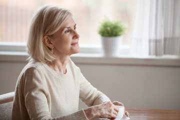 Thoughtful senior woman drinking coffee or tea dreaming about something, dreamy aged female...